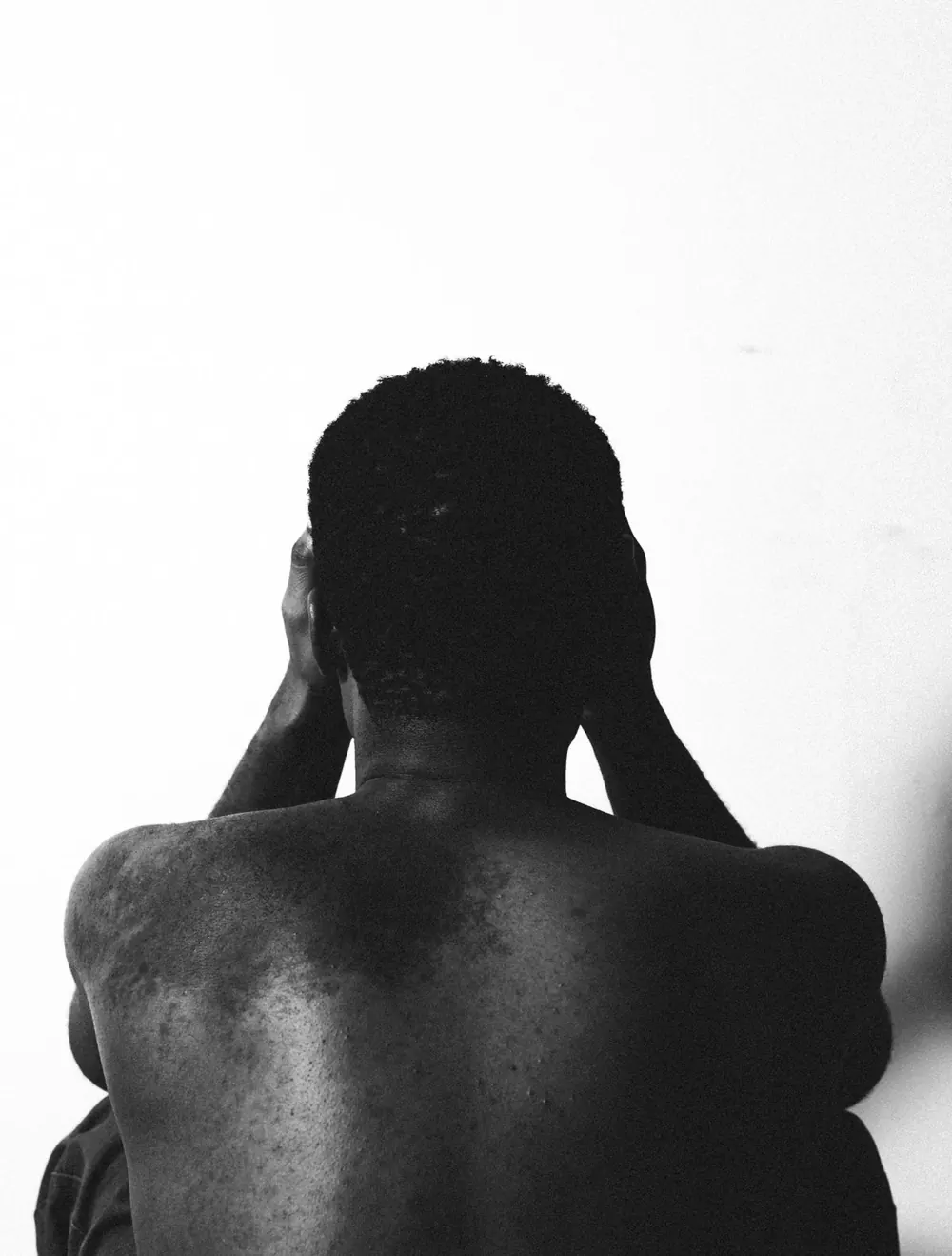 Black-and-white photo of a man seen from behind, bare back, hands resting on his head
