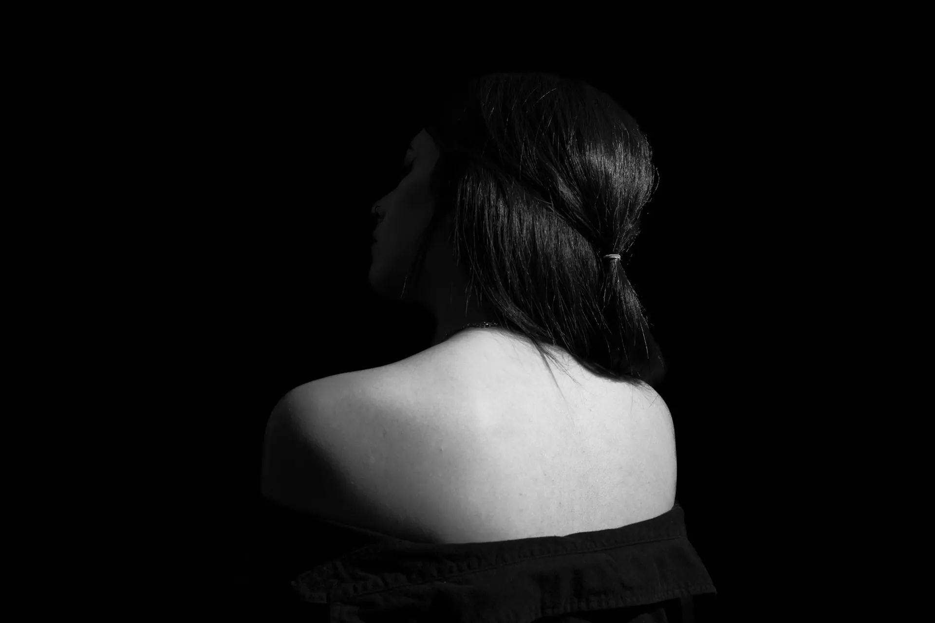 Black-and-white portrait of a woman seen from behind, bare shoulders in soft light against a dark background